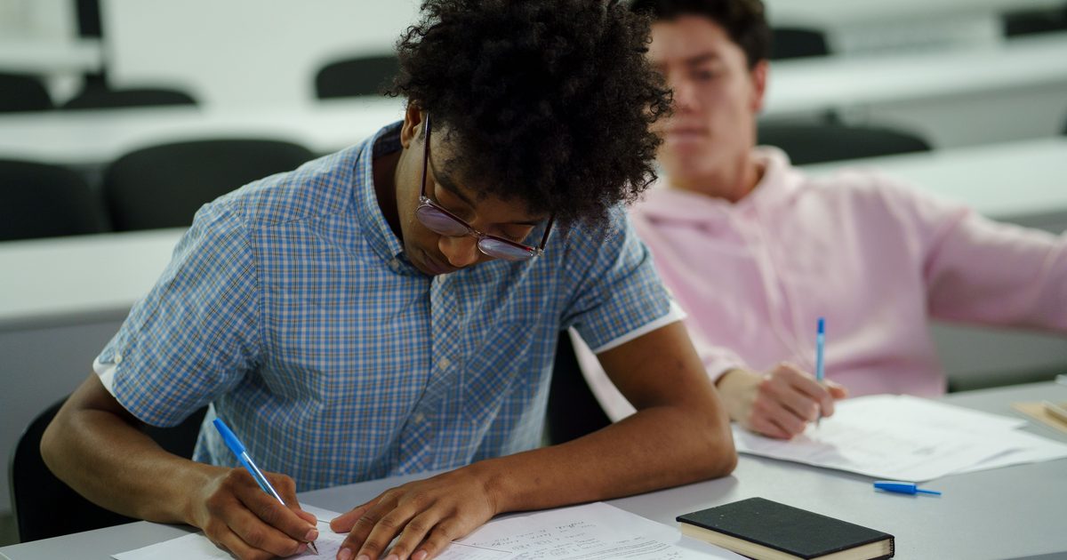 Two students sit at classroom desks writing on exam papers with pens.