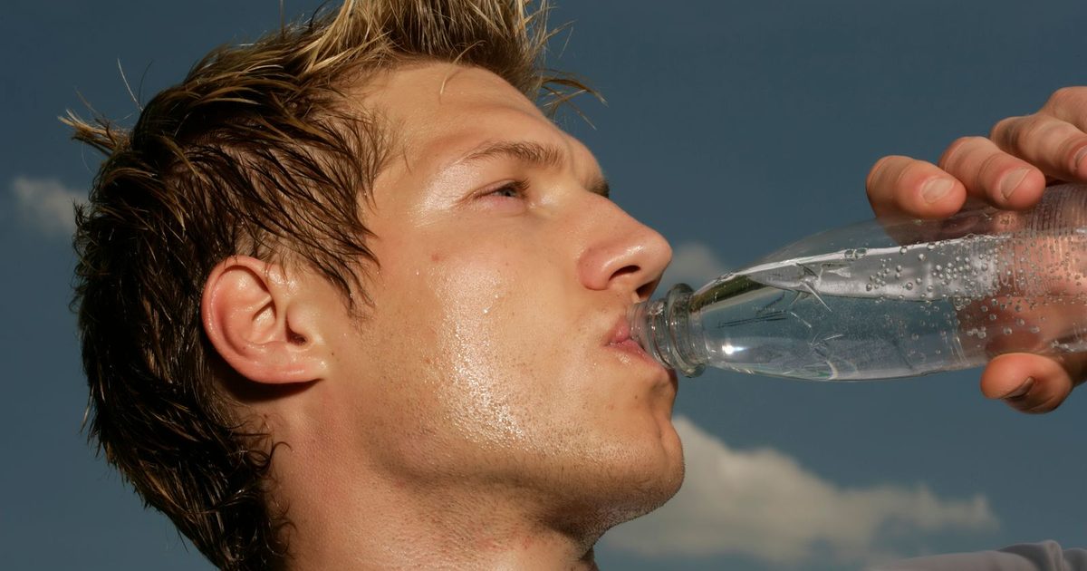 Close-up of a sweaty athletic man drinking water from a bottle outdoors under a blue sky, cooling off after exercise.