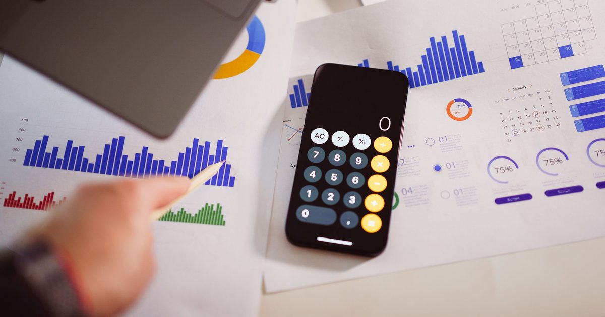 A smartphone showing a calculator app resting on printed sheets of colourful bar charts and pie charts beside a January calendar, with a hand holding a pencil.